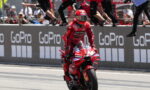 Red-suited motorcycle racer on a red Ducati, riding past GoPro banners with cheering spectators in the background.