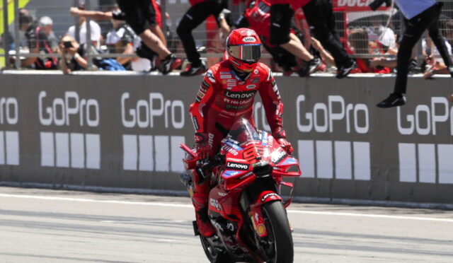 Red-suited motorcycle racer on a red Ducati, riding past GoPro banners with cheering spectators in the background.