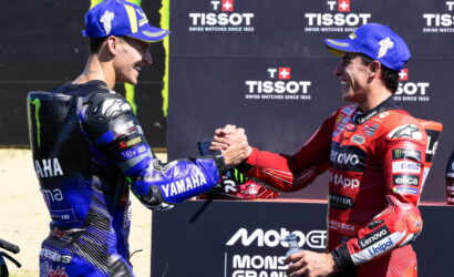 Two motorcycle racers in blue Yamaha and red Ducati suits shake hands on a podium, smiling after a race, with sponsor banners in the background.
