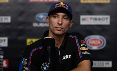 Male race driver at a press conference wearing a Red Bull cap and black team polo, speaking into a microphone with sponsor logos in the backdrop.