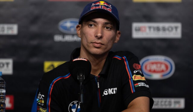 Male race driver at a press conference wearing a Red Bull cap and black team polo, speaking into a microphone with sponsor logos in the backdrop.