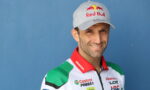 Portrait of a male race car driver wearing a gray Red Bull cap and a white, red, and green sponsor racing suit against a blue background.