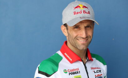 Portrait of a male race car driver wearing a gray Red Bull cap and a white, red, and green sponsor racing suit against a blue background.