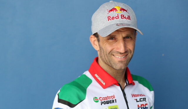 Portrait of a male race car driver wearing a gray Red Bull cap and a white, red, and green sponsor racing suit against a blue background.