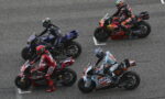 Top-down view of four riders in colorful leathers lined up on racing motorcycles at a grid on the track, ready to start race action.