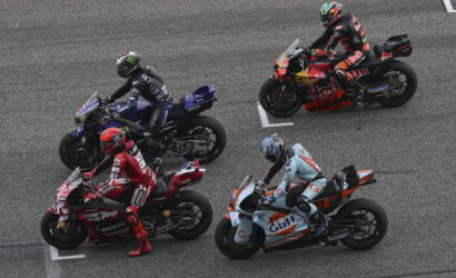 Top-down view of four riders in colorful leathers lined up on racing motorcycles at a grid on the track, ready to start race action.