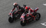 Two motorcycle racers lean into a curve, riding side by side on a gray race track and nearing the turn edges in a tight duel on sport bikes.