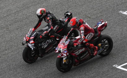 Two motorcycle racers lean into a curve, riding side by side on a gray race track and nearing the turn edges in a tight duel on sport bikes.