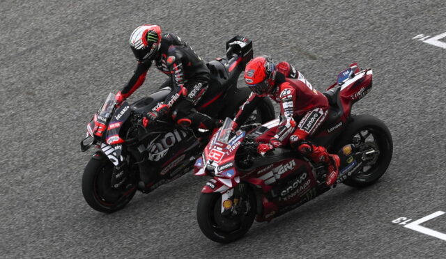 Two motorcycle racers lean into a curve, riding side by side on a gray race track and nearing the turn edges in a tight duel on sport bikes.