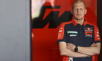 Man with short hair wearing a Red Bull KTM Factory Racing polo, arms crossed, standing against a red backdrop with a dark logo behind him.