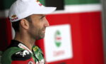 Side profile of a male race car driver wearing a white Red Bull cap and green racing suit, standing in the paddock area