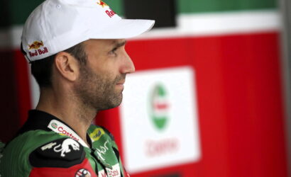 Side profile of a male race car driver wearing a white Red Bull cap and green racing suit, standing in the paddock area