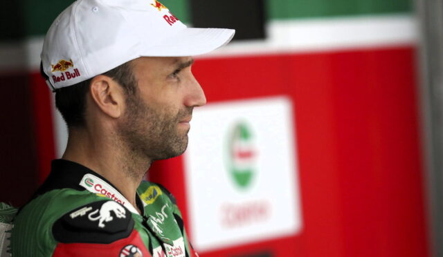 Side profile of a male race car driver wearing a white Red Bull cap and green racing suit, standing in the paddock area