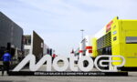 Large white MotoGP sign with Estrella Galicia 0,0 Grand Prix of Spain 2026 text, set in a race paddock with trailers and a bright yellow service bus nearby
