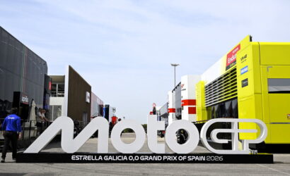 Large white MotoGP sign with Estrella Galicia 0,0 Grand Prix of Spain 2026 text, set in a race paddock with trailers and a bright yellow service bus nearby