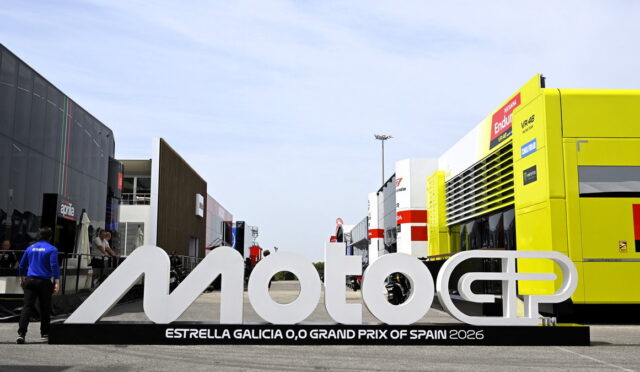 Large white MotoGP sign with Estrella Galicia 0,0 Grand Prix of Spain 2026 text, set in a race paddock with trailers and a bright yellow service bus nearby