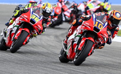 Motorcycle race: red racing bikes lean into a curve with racers in full gear, numbers 10 and 36 visible at the front.