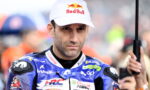 Motorcycle racer in blue racing suit with sponsor logos, wearing a Red Bull cap, holding a black umbrella.