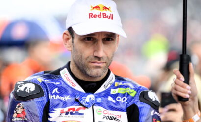 Motorcycle racer in blue racing suit with sponsor logos, wearing a Red Bull cap, holding a black umbrella.