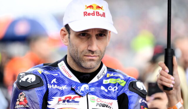 Motorcycle racer in blue racing suit with sponsor logos, wearing a Red Bull cap, holding a black umbrella.