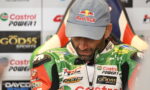 Racer in a light gray Red Bull cap looking down, wearing a green Castrol Power1 racing suit with sponsor patches in a press area.