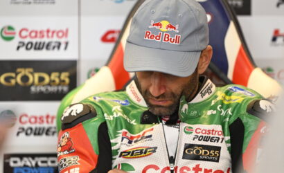Racer in a light gray Red Bull cap looking down, wearing a green Castrol Power1 racing suit with sponsor patches in a press area.