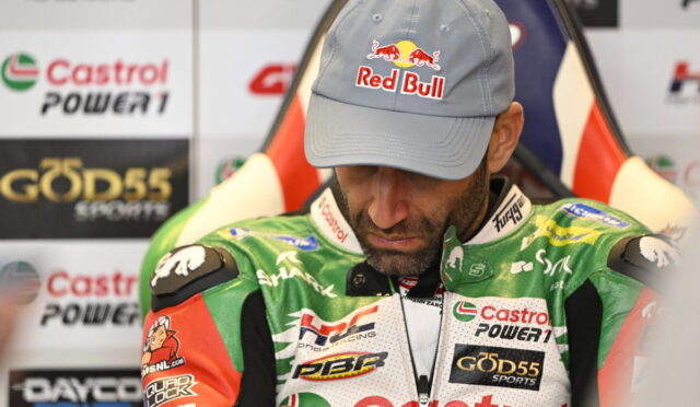 Racer in a light gray Red Bull cap looking down, wearing a green Castrol Power1 racing suit with sponsor patches in a press area.