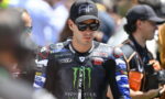 MotoGP rider in black Monster Yamaha suit wearing sunglasses and a cap, standing among fans at a race event.
