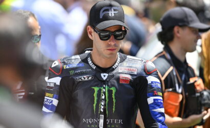 MotoGP rider in black Monster Yamaha suit wearing sunglasses and a cap, standing among fans at a race event.