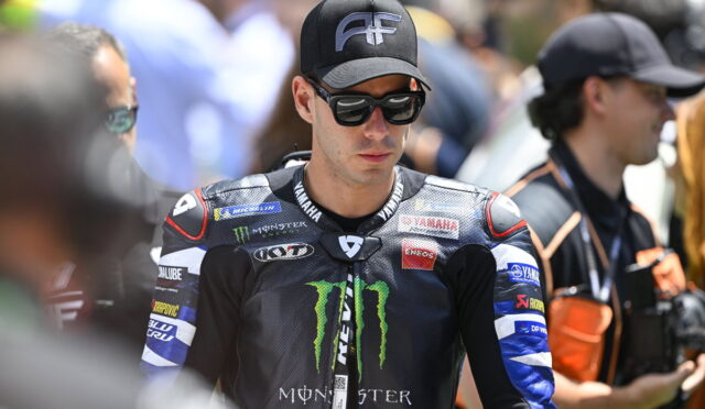 MotoGP rider in black Monster Yamaha suit wearing sunglasses and a cap, standing among fans at a race event.