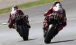 Two motorcycle racers in red leathers leaning into a turn on a racetrack, seen from behind.