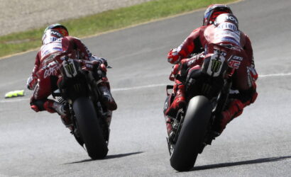 Two motorcycle racers in red leathers leaning into a turn on a racetrack, seen from behind.
