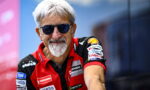 Older man with gray hair and beard wearing dark sunglasses, in a red racing team shirt with sponsor logos, smiling at the camera.