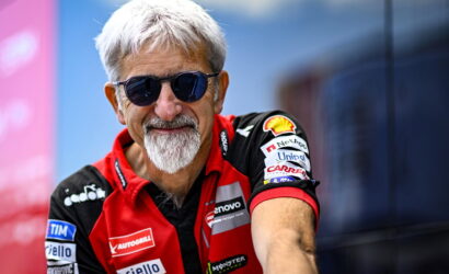 Older man with gray hair and beard wearing dark sunglasses, in a red racing team shirt with sponsor logos, smiling at the camera.