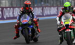 Two motorcycle racers wheel-to-wheel on a race track, front bike number 20 wearing blue and red with Monster energy graphics.