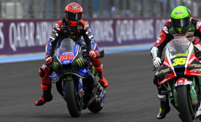 Two motorcycle racers wheel-to-wheel on a race track, front bike number 20 wearing blue and red with Monster energy graphics.