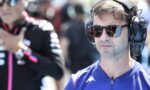 Man wearing large over-ear headset and sunglasses at a motorsport event, focused on something off-camera.
