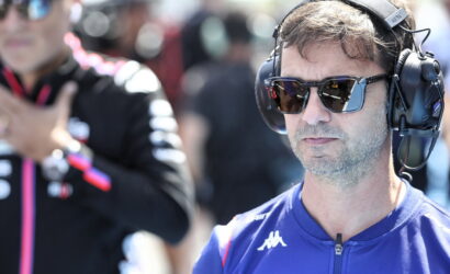 Man wearing large over-ear headset and sunglasses at a motorsport event, focused on something off-camera.