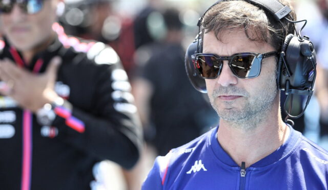 Man wearing large over-ear headset and sunglasses at a motorsport event, focused on something off-camera.