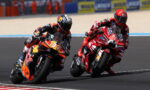 Two motorcycle racers in red and black leathers lean into a turn on a racetrack, speeding side by side.