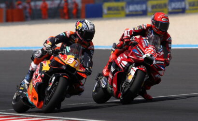 Two motorcycle racers in red and black leathers lean into a turn on a racetrack, speeding side by side.