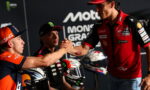 Rider in orange and navy cap shakes hands with a Ducati rider on stage, while a teammate watches, with helmets nearby.