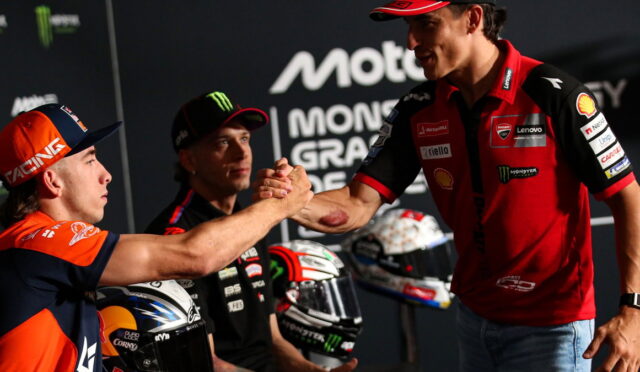 Rider in orange and navy cap shakes hands with a Ducati rider on stage, while a teammate watches, with helmets nearby.