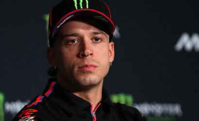 Athlete wearing a black cap with red trim and Monster logo at a press conference with a dark backdrop.