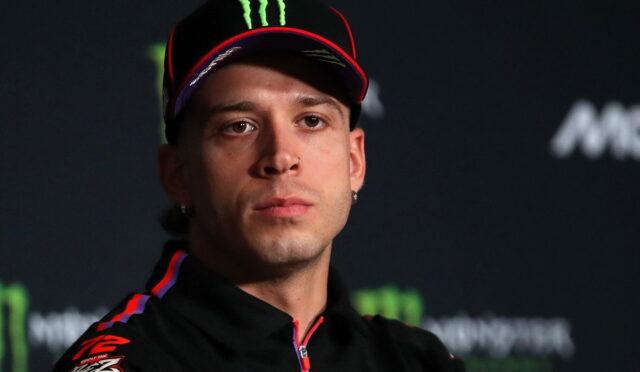 Athlete wearing a black cap with red trim and Monster logo at a press conference with a dark backdrop.