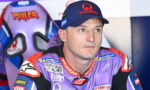 MotoGP rider in a purple Pramac racing suit and cap, seated in the pit area and looking to the side.