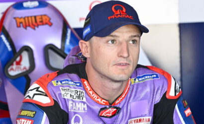 MotoGP rider in a purple Pramac racing suit and cap, seated in the pit area and looking to the side.
