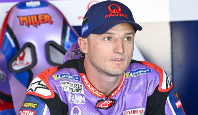 MotoGP rider in a purple Pramac racing suit and cap, seated in the pit area and looking to the side.