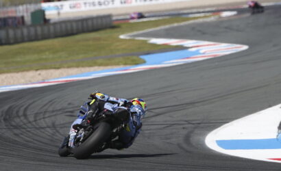 Motorcycle racer in a blue riding suit leaning into a sharp turn on a racetrack, with barriers and grass in the background.