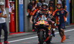 MotoGP rider in full gear on an orange KTM bike in the pit lane, flanked by crew in orange and navy.